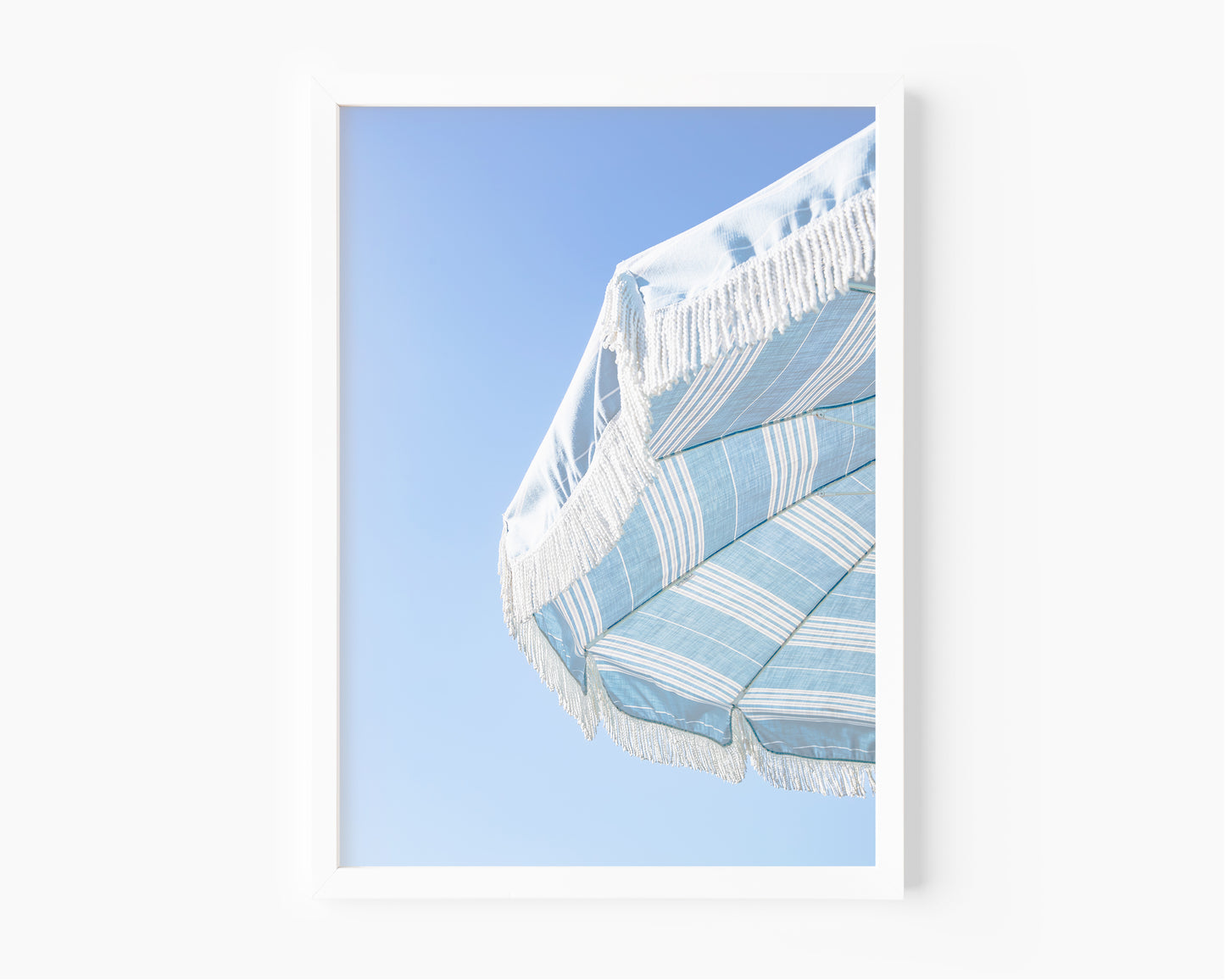 Blue and white striped beach umbrella with fringe set against a clear sky in a minimalist vertical beach photograph.