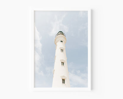 White lighthouse against a blue sky and soft clouds in Aruba – beach photography print for coastal wall decor.