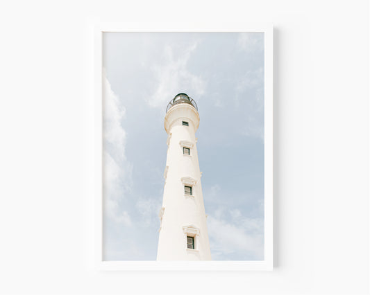 White lighthouse against a blue sky and soft clouds in Aruba – beach photography print for coastal wall decor.