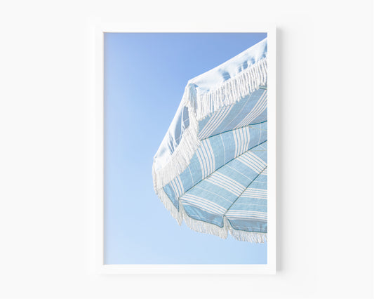 Blue and white striped beach umbrella with fringe set against a clear sky in a minimalist vertical beach photograph.