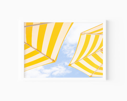 Yellow and white striped beach umbrellas set against a bright blue sky with scattered clouds; minimalist beach photography evoking a sunny Amalfi Coast vibe.